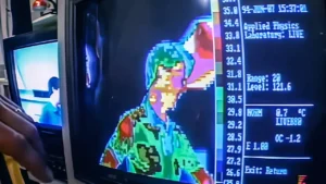 Scientist pointing an infrared camera at objects to demonstrate invisible thermal radiation with colour thermal image visible on screen
