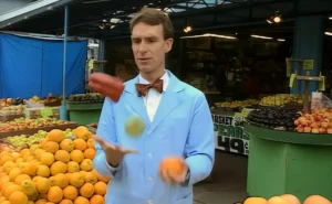 Bill Nye wearing a light blue lab coat and bow tie juggling a red pepper, an orange, and a green apple at a fruit market stall to demonstrate colour reflection.