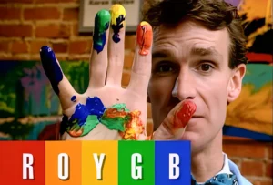 Bill Nye holding up a hand covered in colourful paint above graphic blocks spelling out the letters R O Y G B.