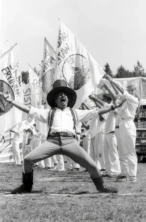 Paul Zaloom in a wide brimmed hat, arms flung out and mouth open, announcing the Bread and Puppet Domestic Resurrection Circus in a Vermont field
