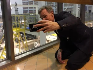 Bill Nye crouching at a viewing window, holding up a phone to photograph the James Webb Space Telescope under construction in the cleanroom beyond