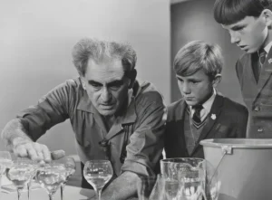 Julius Sumner Miller reaching across a table of water-filled wine glasses while two schoolboys in blazers watch closely