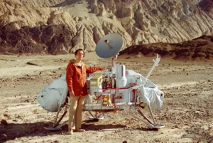 Carl Sagan in an orange jacket standing beside a full-size Viking lander model in the desert at Death Valley, California
