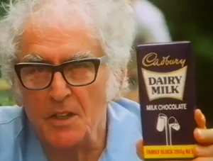 Julius Sumner Miller in later life, in thick black-framed glasses, holding up a family block of Cadbury Dairy Milk to camera