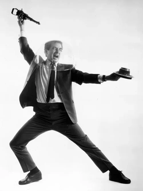 Paul Zaloom in a suit and a clear salad spinner helmet, legs braced apart, holding a small gun in one hand and a cake in the other