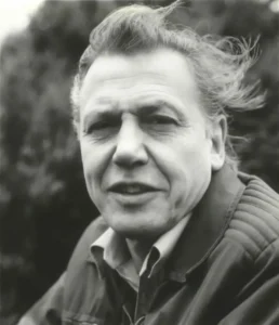 Black and white press photograph of David Attenborough in 1982, outdoors in a field jacket, hair blown across his forehead