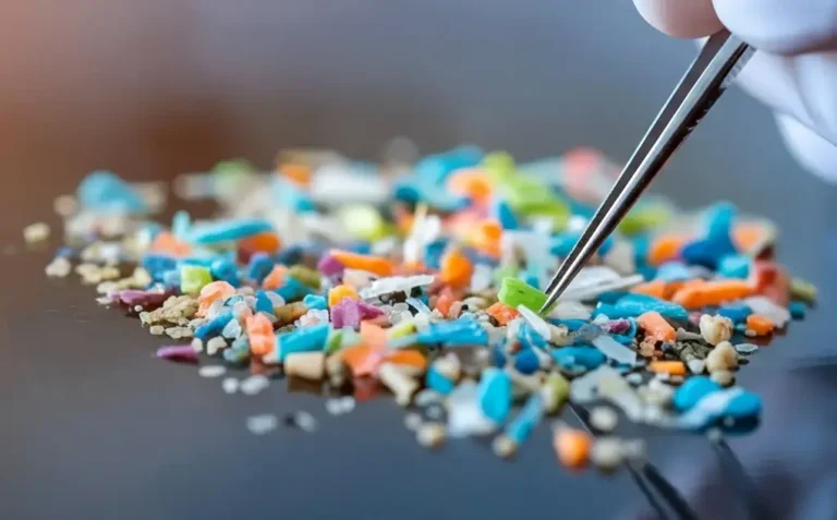 Close-up of colorful microplastic fragments mixed with sand particles on a dark surface, with tweezers picking up one piece.
