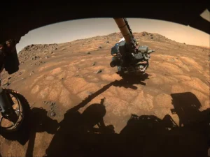 NASA's Perseverance rover extends its robotic arm over Martian rocks, showing the turret containing scientific instruments like SHERLOC and PIXL.