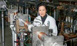 A highly detailed, digitally colorized archival photograph of Dr. Chien-Shiung Wu in a laboratory setting. She is wearing a white lab coat over a dark collar, smiling warmly and directly at the camera. She is surrounded by complex experimental physics equipment, including vacuum lines, metal tubing, and large coils.