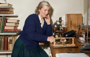 Dorothy Hodgkin sitting at a laboratory desk in 1964 next to a microscope and a molecular crystal structure model.
