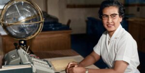 A photograph of Katherine Johnson, an African American woman, seated at a desk at NASA’s Langley Research Center. She is wearing a white dress and glasses, with her hands resting on papers covered in mathematical charts and calculations. Behind her is a celestial globe used for tracking orbits. Digital restoration and colorization by Seriously Scientific.