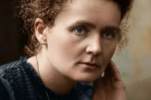 A highly detailed, digitally colorized close-up portrait of scientist Marie Curie. She looks directly at the camera with a serious and focused expression while resting her cheek against her hand. She has curly brown hair and wears a dark blue dress with textured pleats.