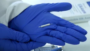 A close-up view of a scientist's hands in blue gloves holding the tiny, tube-shaped SilkAxons device.