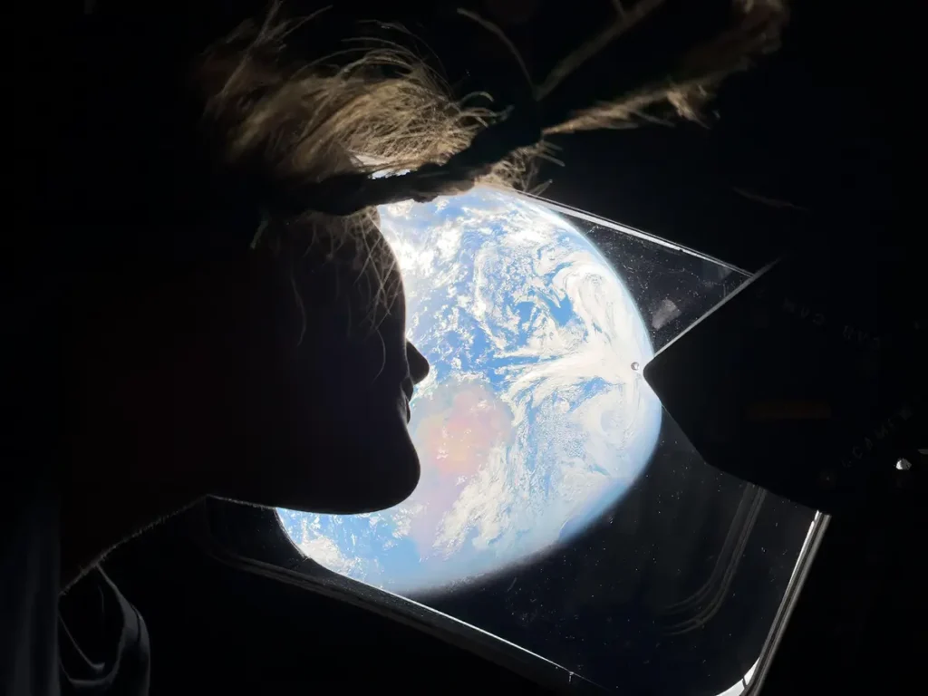A silhouetted profile of NASA astronaut Christina Koch looking out of a window on the Orion spacecraft. Outside the bright window, the blue and white marble of Earth is clearly visible against the blackness of space.