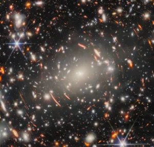 A dense galaxy cluster surrounded by the glowing warped arcs of distant lensed galaxies.