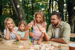 Mother, father and two young girls sitting at an outdoor wooden table, stacking colourful wooden blocks into a tower, with green trees behind them