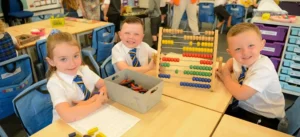 Three young children in blue and white school uniforms with striped ties, sitting at classroom tables with an abacus and a box of crayons on their first day at primary school