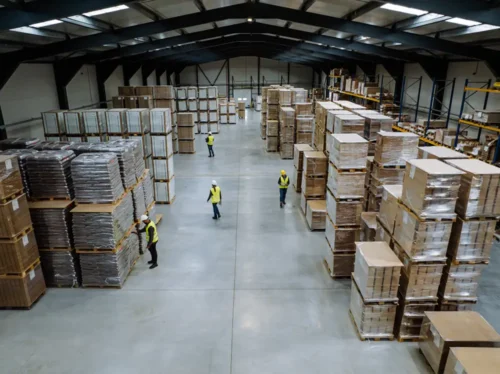 A large industrial warehouse filled with stacked boxes on pallets, with supervisors managing the floor.