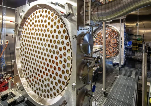 A large circular array of several hundred gold coloured photomultiplier tubes set into a white disc, standing open in a laboratory beside a second array wired with copper cabling.